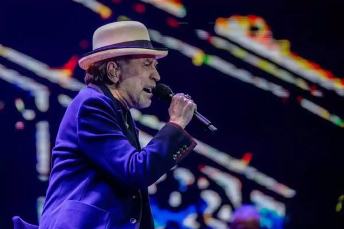 Hombre mayor con sombrero y saco azul cantando en un escenario con luces de colores al fondo