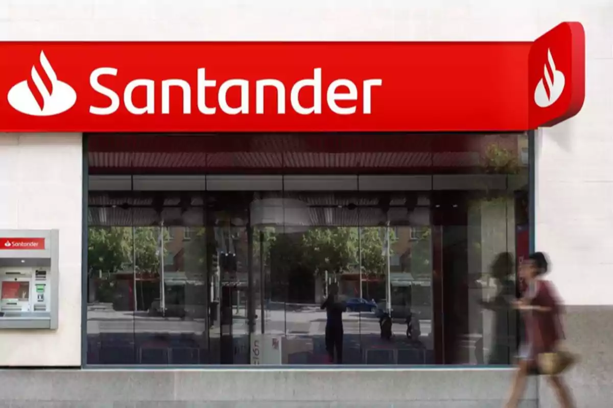 Fachada de una sucursal bancaria de Santander con un cajero automático y una persona caminando frente al edificio Fachada de una sucursal bancaria de Santander con un cajero automático y una persona caminando frente al edificio