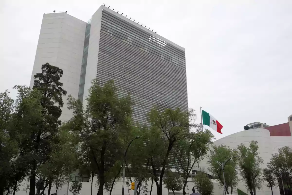 Edificio moderno de gran altura rodeado de árboles con una bandera de México ondeando al frente Edificio moderno de gran altura rodeado de árboles con una bandera de México ondeando al frente