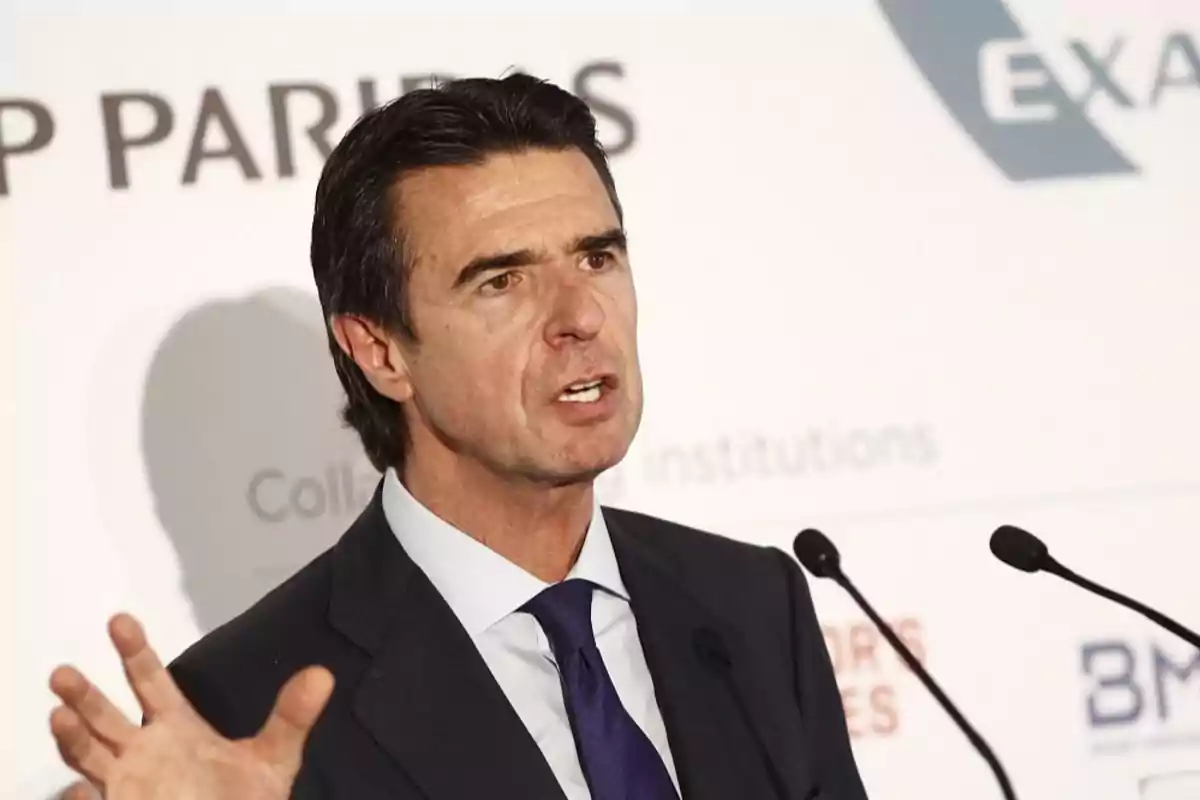 Hombre de cabello oscuro con traje y corbata hablando frente a dos micrófonos en un evento profesional Hombre de cabello oscuro con traje y corbata hablando frente a dos micrófonos en un evento profesional