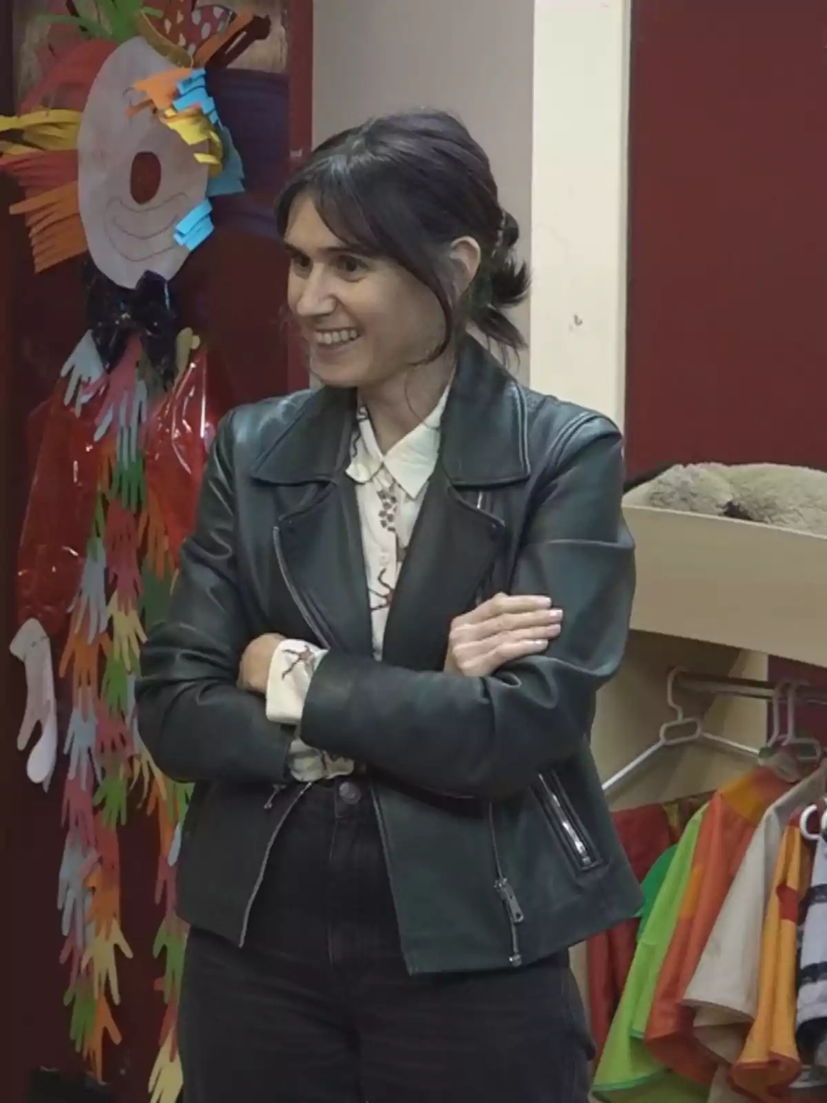 Mujer sonriente de cabello oscuro y chaqueta de cuero negro con los brazos cruzados en una habitación decorada con disfraces y un payaso hecho de papel en la pared Mujer sonriente de cabello oscuro y chaqueta de cuero negro con los brazos cruzados en una habitación decorada con disfraces y un payaso hecho de papel en la pared