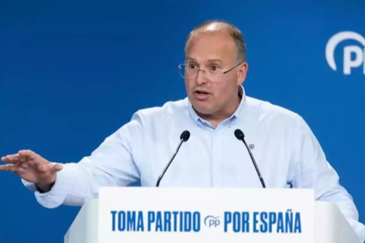 Hombre con gafas y camisa clara hablando en un atril con el texto Toma partido por España sobre un fondo azul Hombre con gafas y camisa clara hablando en un atril con el texto Toma partido por España sobre un fondo azul