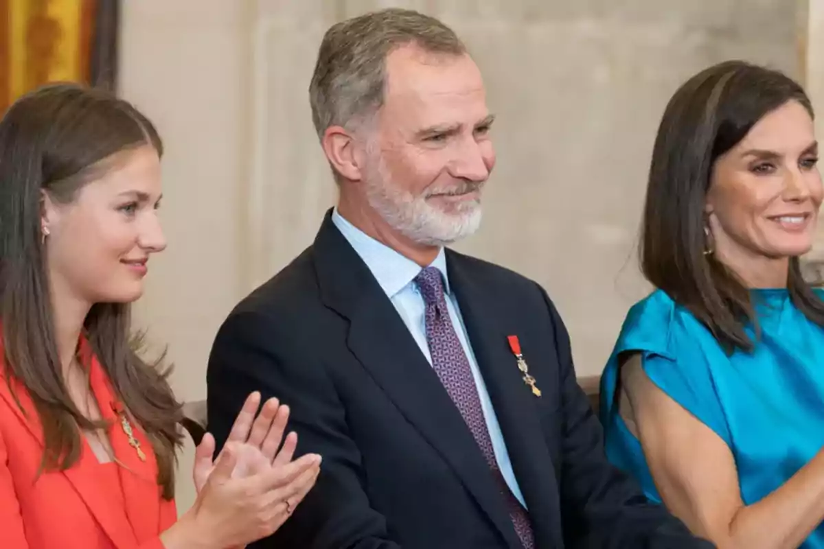 Three elegantly dressed people applaud and smile at a formal event.