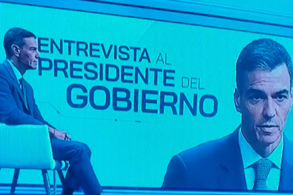 Hombre con traje sentado en un plató de televisión junto a una pantalla azul que muestra el texto entrevista al presidente del gobierno y su retrato en grande Hombre con traje sentado en un plató de televisión junto a una pantalla azul que muestra el texto entrevista al presidente del gobierno y su retrato en grande