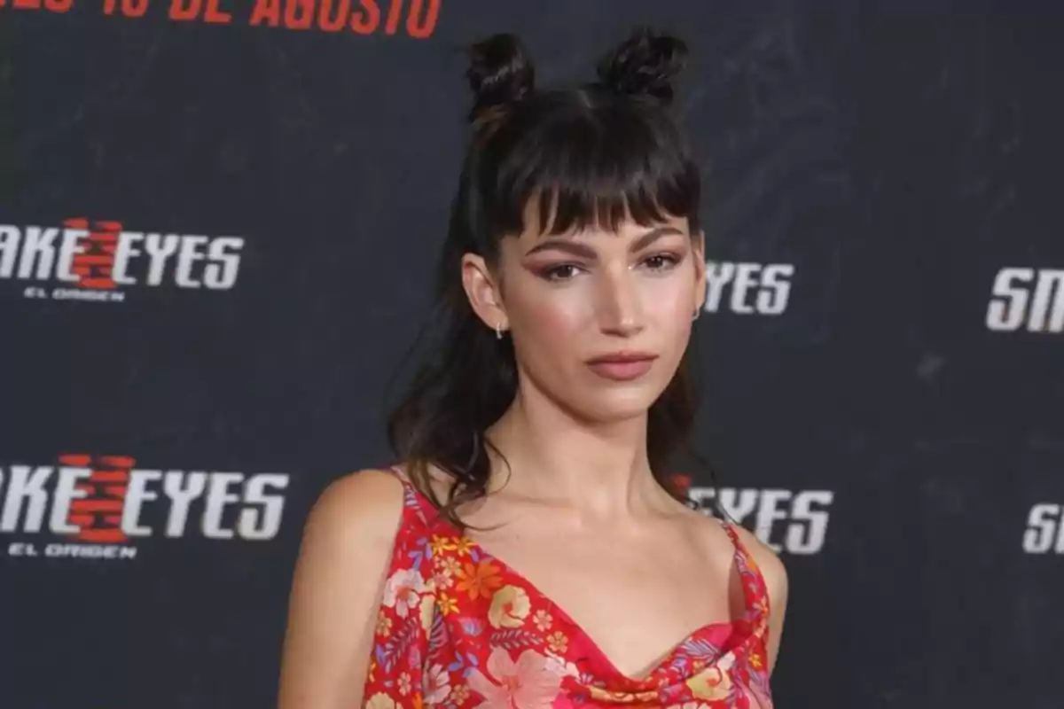 Mujer con peinado de dos moños y flequillo usando un vestido rojo con estampado floral posando frente a un fondo promocional de una película Mujer con peinado de dos moños y flequillo usando un vestido rojo con estampado floral posando frente a un fondo promocional de una película