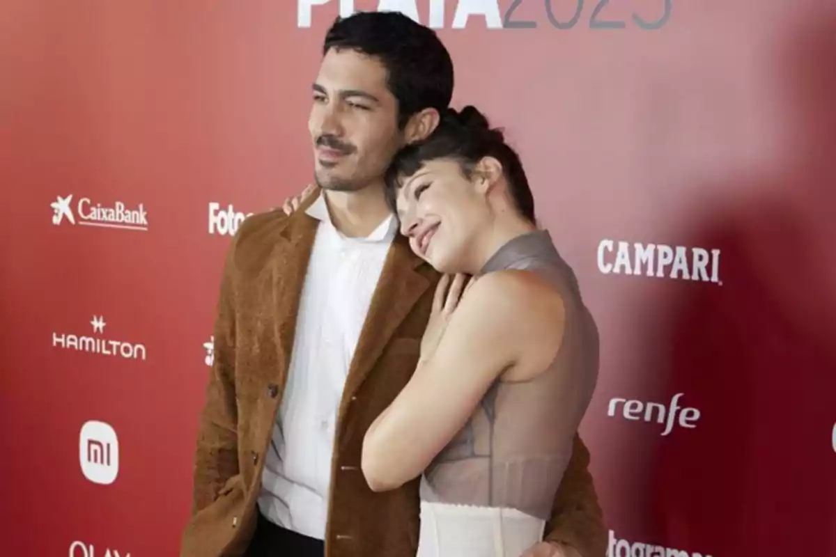 Pareja posando sonriente en una alfombra roja frente a un fondo con logotipos de marcas y letras blancas Pareja posando sonriente en una alfombra roja frente a un fondo con logotipos de marcas y letras blancas
