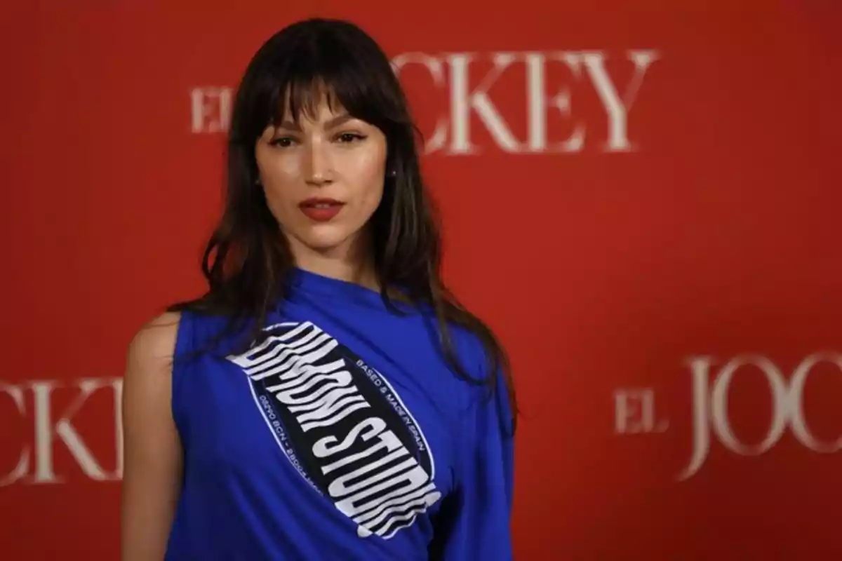 Mujer de cabello castaño y flequillo usando una blusa azul con letras blancas posando frente a un fondo rojo con texto borroso