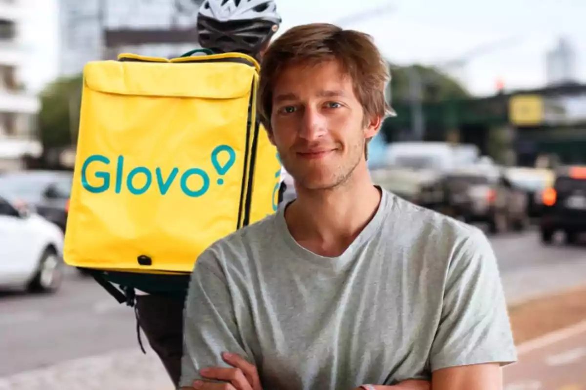 Hombre joven de cabello castaño claro con camiseta gris sonriendo frente a un repartidor de Glovo en bicicleta en una calle urbana