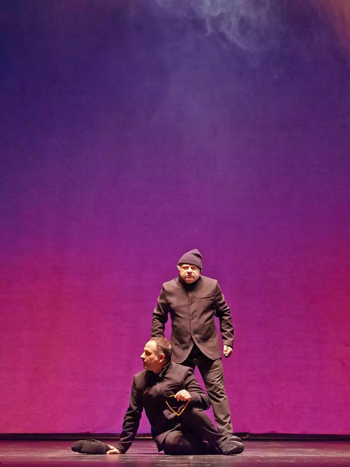 Dos actores vestidos de negro interpretan una escena teatral sobre un escenario iluminado con fondo morado Dos actores vestidos de negro interpretan una escena teatral sobre un escenario iluminado con fondo morado