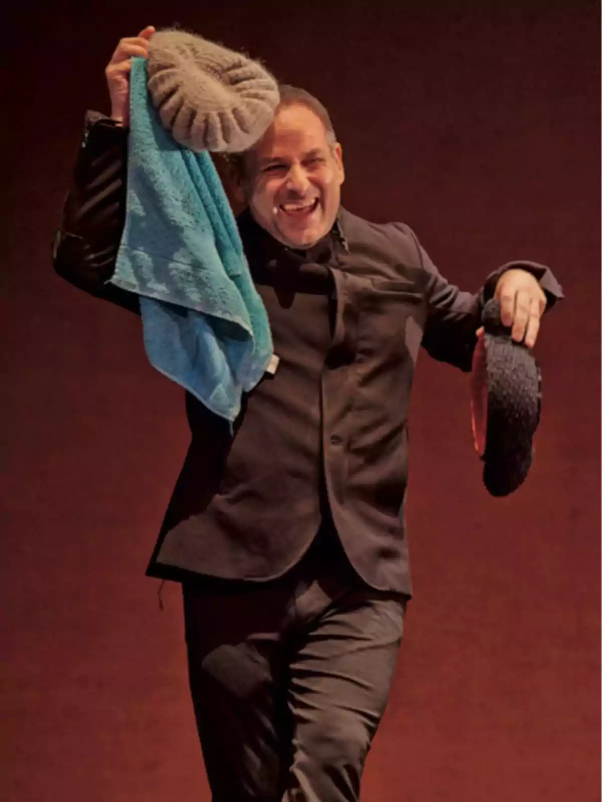 Hombre sonriente vestido de traje oscuro sostiene una boina gris y una toalla azul en una mano y un objeto negro en la otra sobre un fondo marrón Hombre sonriente vestido de traje oscuro sostiene una boina gris y una toalla azul en una mano y un objeto negro en la otra sobre un fondo marrón
