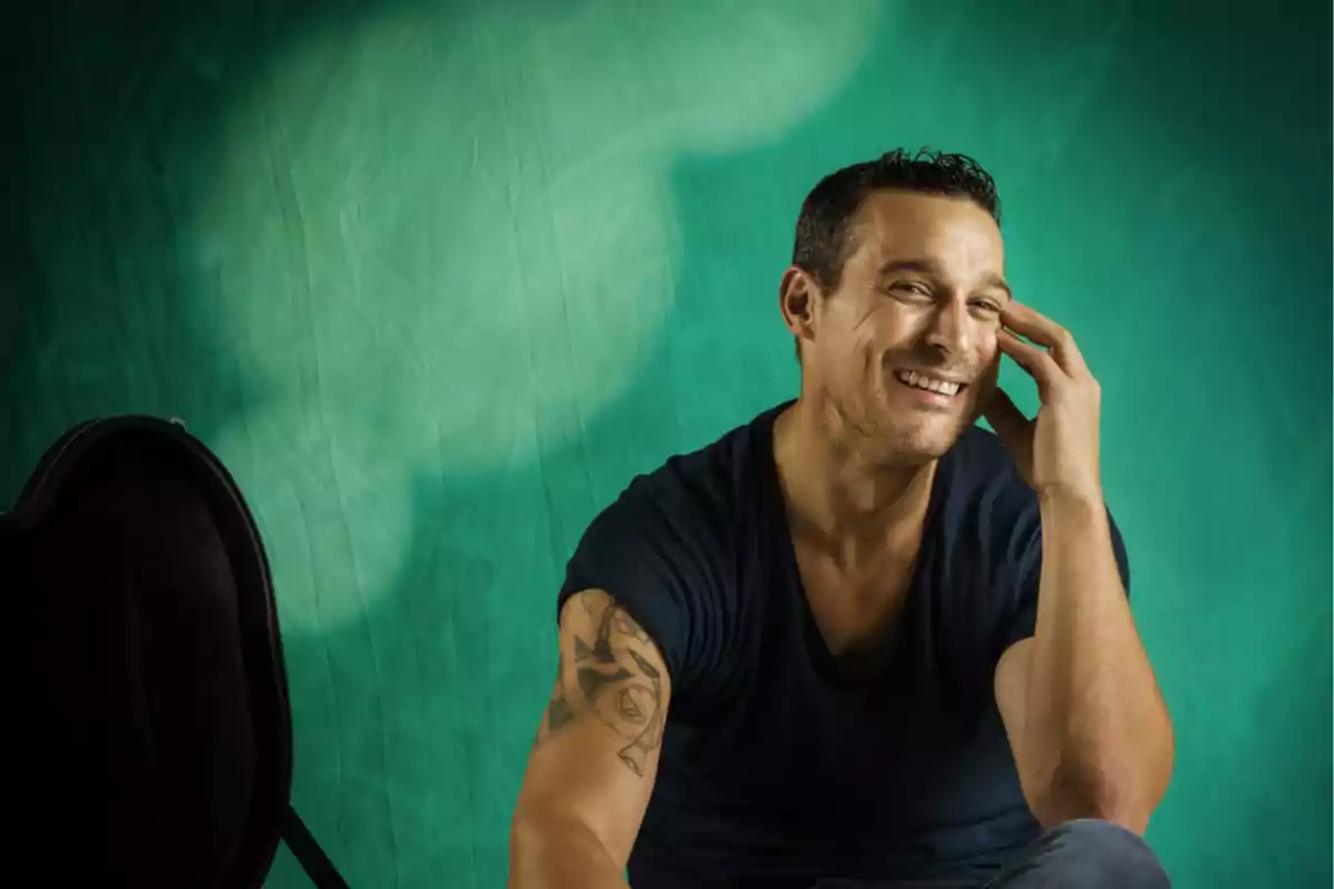 Hombre sonriente con camiseta oscura y tatuaje en el brazo sentado frente a un fondo verde Hombre sonriente con camiseta oscura y tatuaje en el brazo sentado frente a un fondo verde