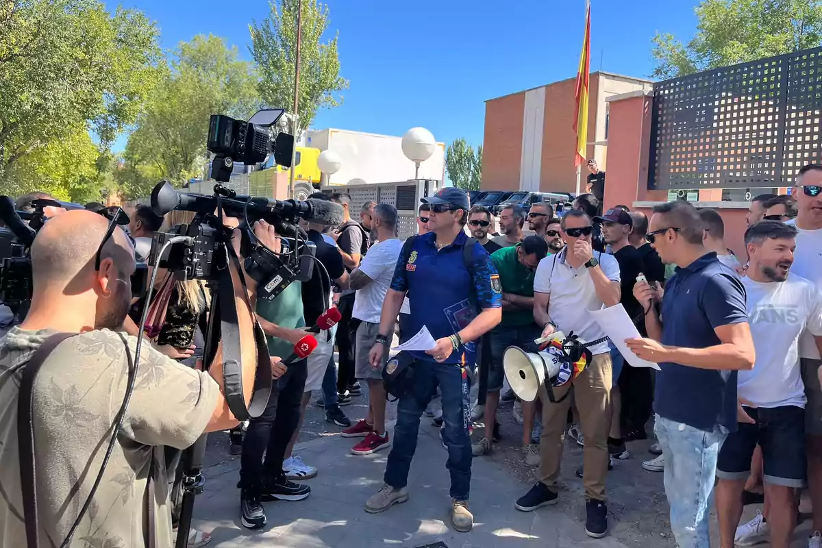 Un grupo de personas se reúne al aire libre frente a cámaras de televisión y periodistas mientras un hombre con uniforme policial sostiene papeles y otro lleva un megáfono en la mano Un grupo de personas se reúne al aire libre frente a cámaras de televisión y periodistas mientras un hombre con uniforme policial sostiene papeles y otro lleva un megáfono en la mano
