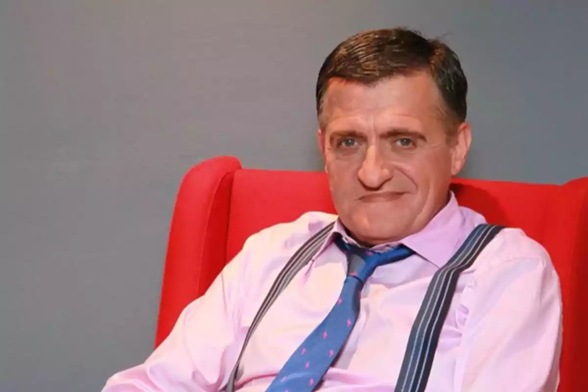 Hombre de mediana edad con camisa rosa y tirantes sentado en un sillón rojo frente a una pared gris Hombre de mediana edad con camisa rosa y tirantes sentado en un sillón rojo frente a una pared gris