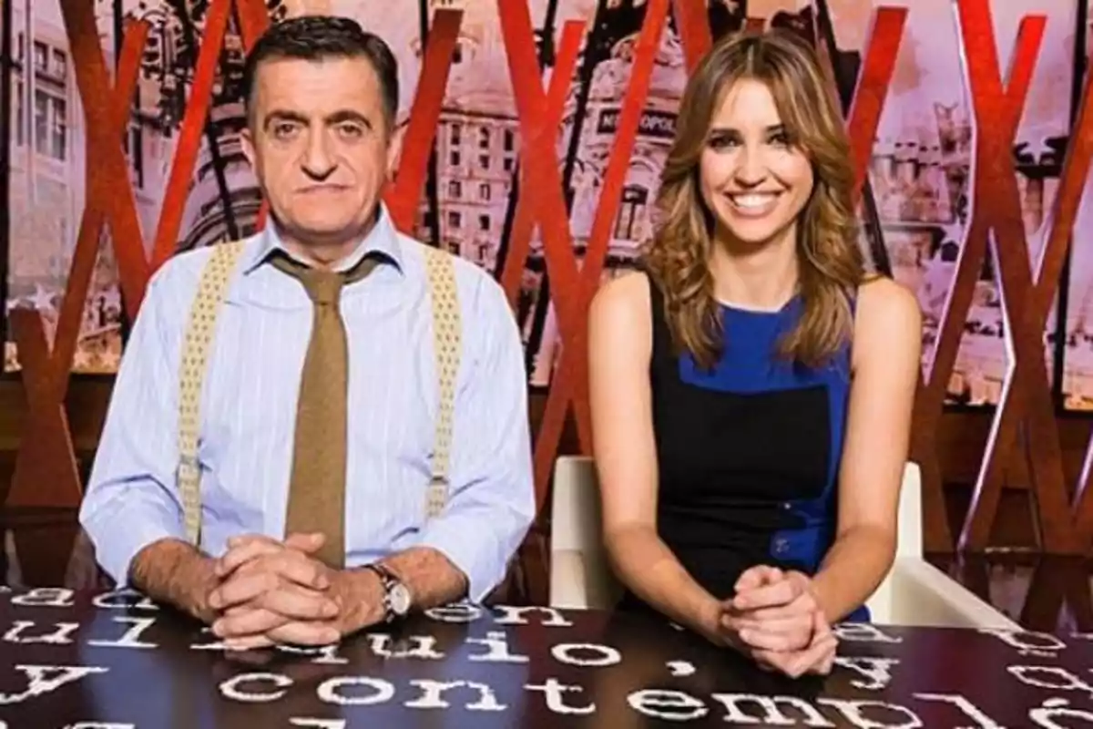 Dos presentadores sentados detrás de una mesa con letras blancas sobre fondo decorativo rojo y urbano Dos presentadores sentados detrás de una mesa con letras blancas sobre fondo decorativo rojo y urbano