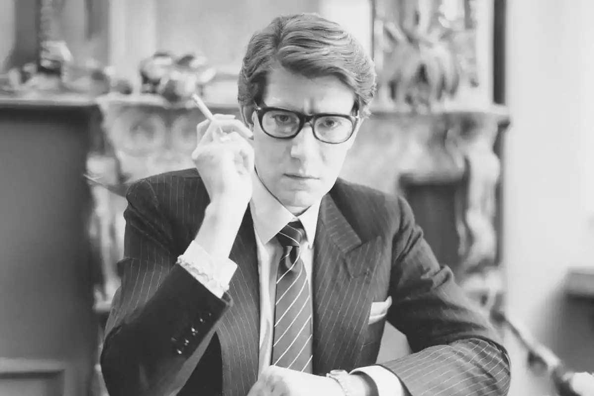 Hombre joven con gafas y traje a rayas sosteniendo un cigarrillo en una mano mientras mira fijamente a la cámara en una habitación elegante