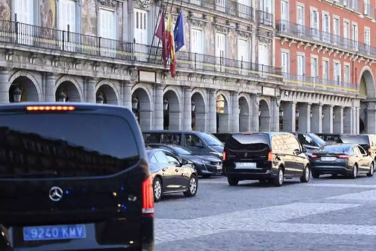 Una fila de vehículos negros circula frente a un edificio histórico con arcos y banderas.
