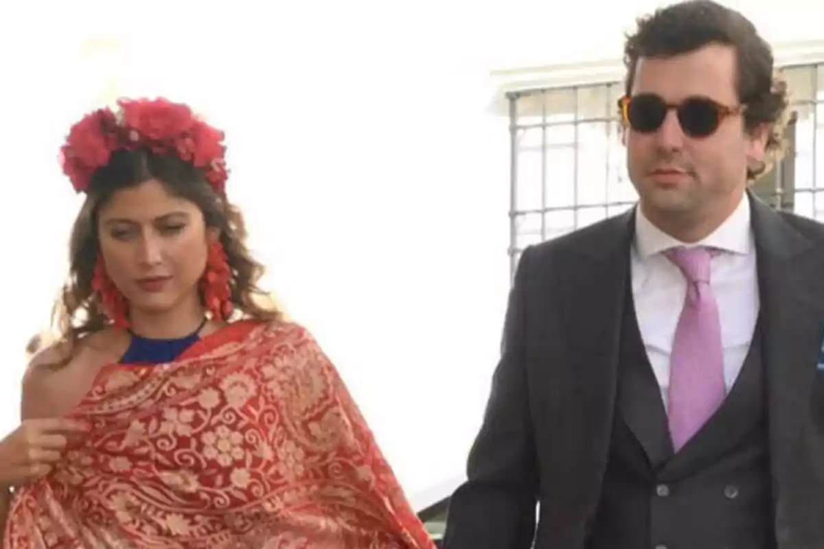 Una mujer con una corona de flores rojas y un chal rojo con patrones dorados camina junto a un hombre con gafas de sol, traje oscuro y corbata rosa.