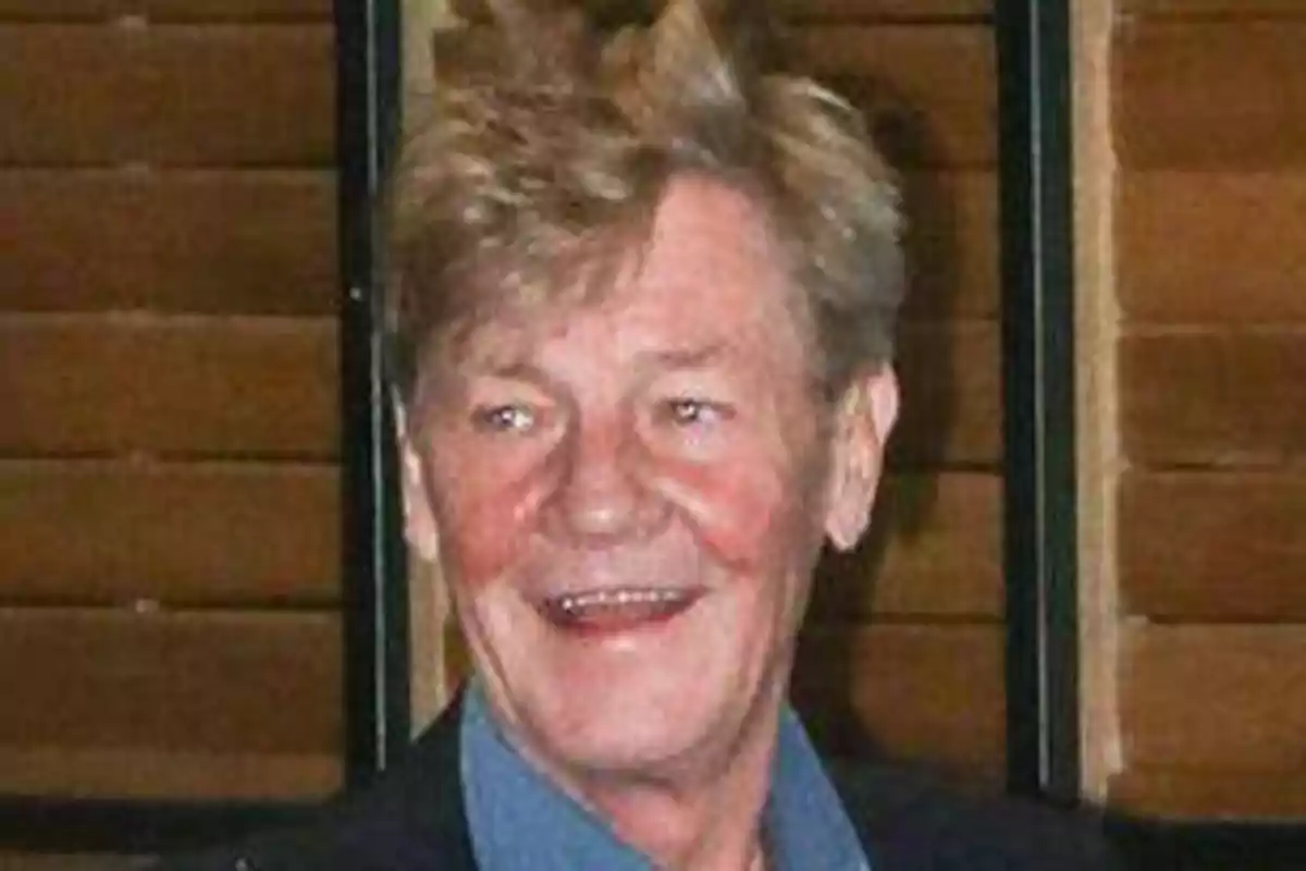 Un hombre mayor con cabello rubio y una expresión sonriente, con un fondo de persianas de madera.