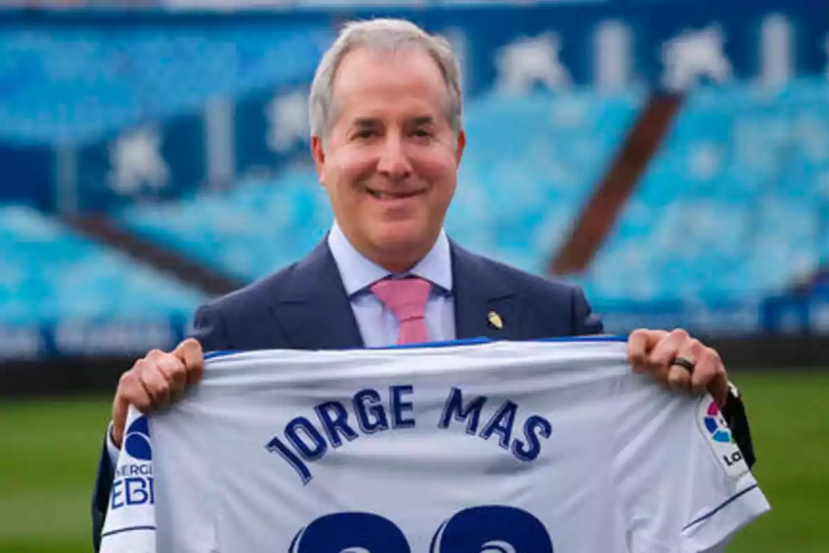 Un hombre sonriente sostiene una camiseta de fútbol con el nombre "Jorge Mas" en un estadio vacío.