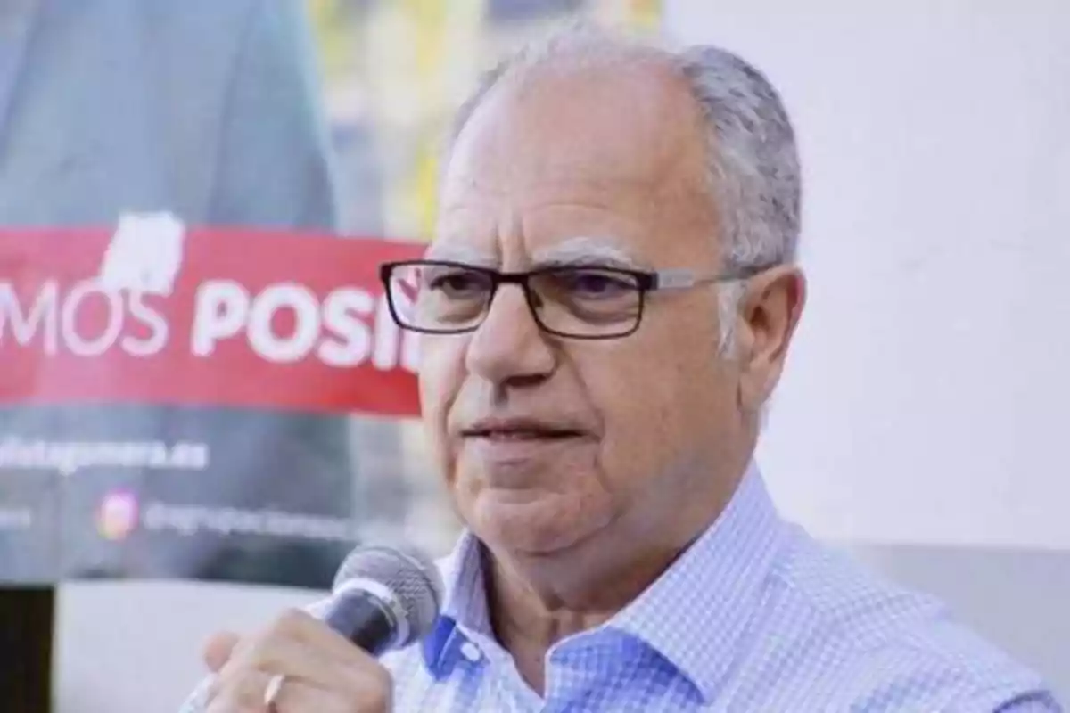 Hombre mayor con gafas hablando en un micrófono frente a un cartel.