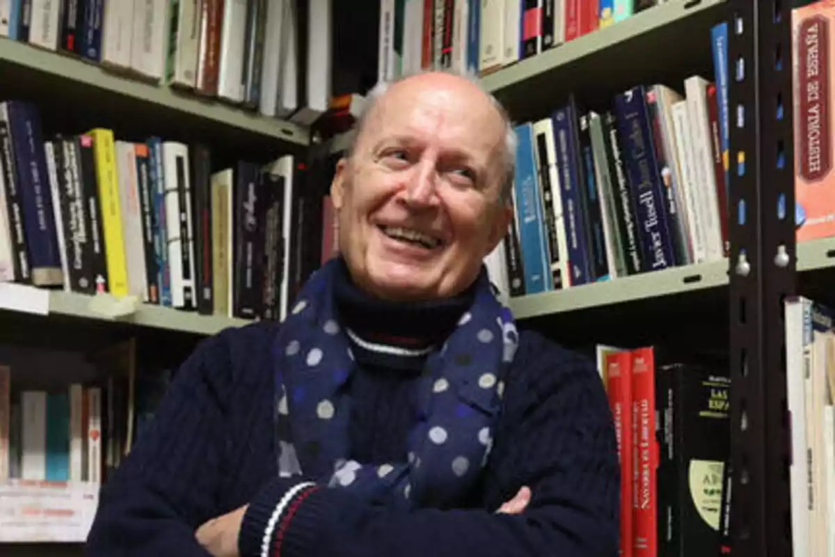 Un hombre sonriente con bufanda azul de lunares, de pie frente a una estantería llena de libros.