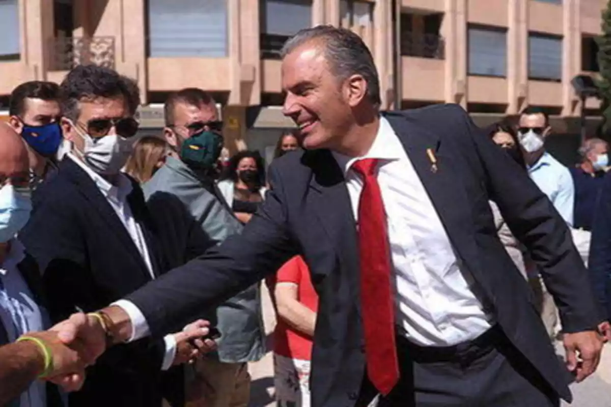 Un hombre con traje y corbata roja estrechando la mano de otra persona en un evento al aire libre, rodeado de varias personas que llevan mascarillas.