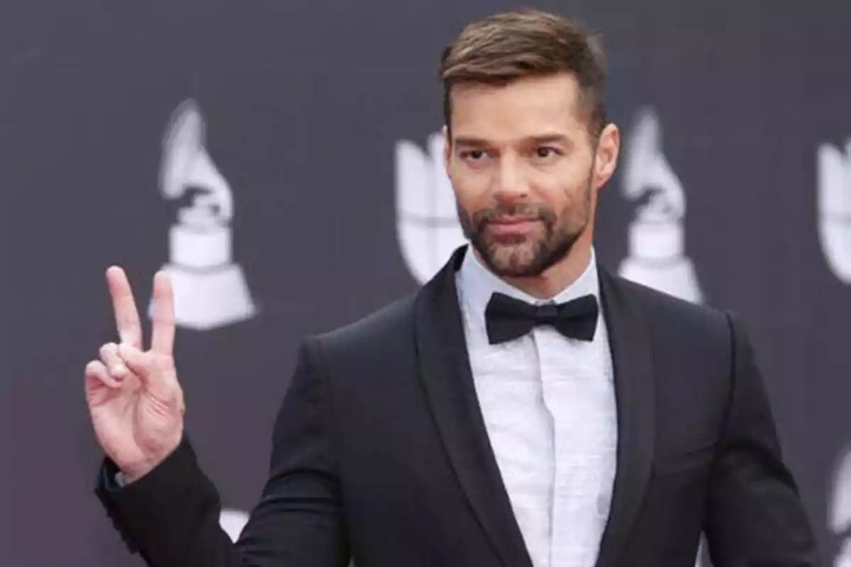 Hombre con barba y traje negro haciendo el signo de la paz con los dedos frente a un fondo con logotipos de premios.