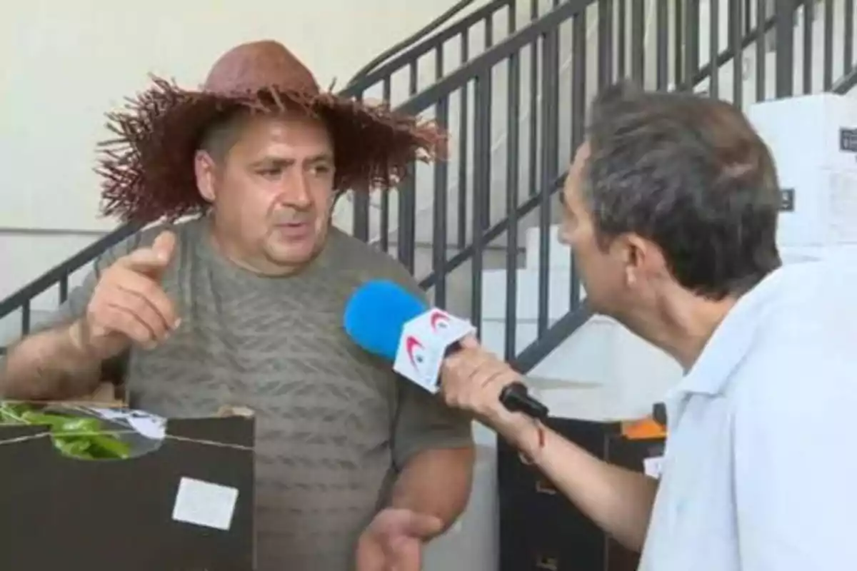 Un hombre con sombrero de paja siendo entrevistado por un reportero con un micrófono azul.