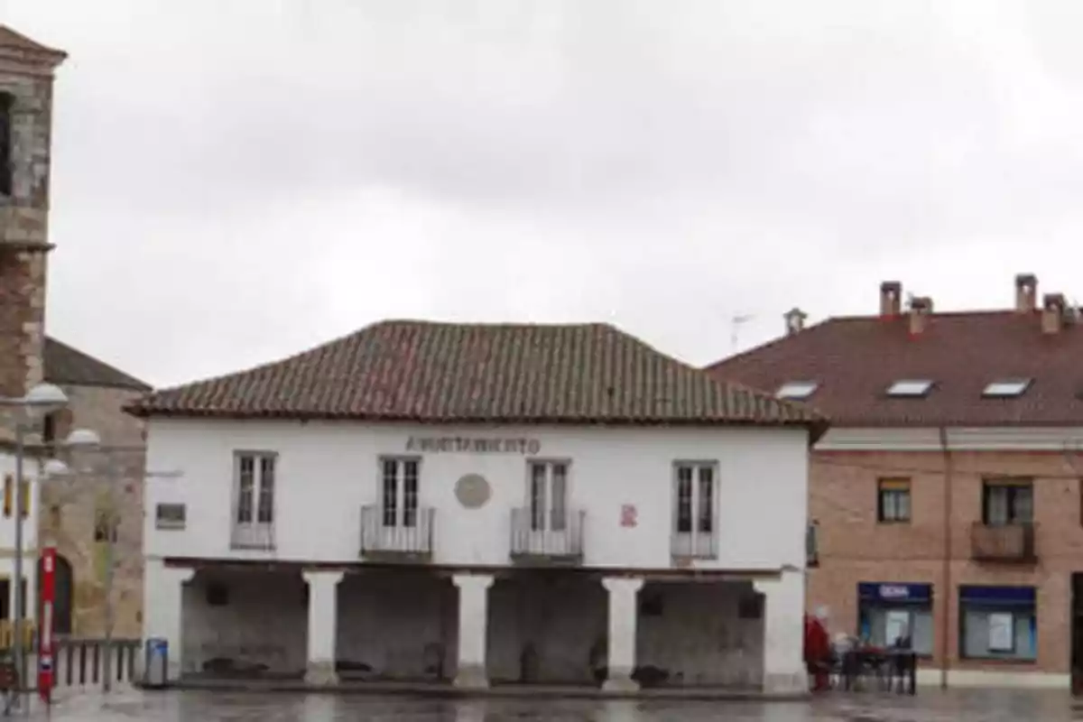 Imagen de un edificio con la palabra "Ayuntamiento" en la fachada, con un techo de tejas y balcones, ubicado en una plaza con suelo mojado y rodeado de otros edificios.