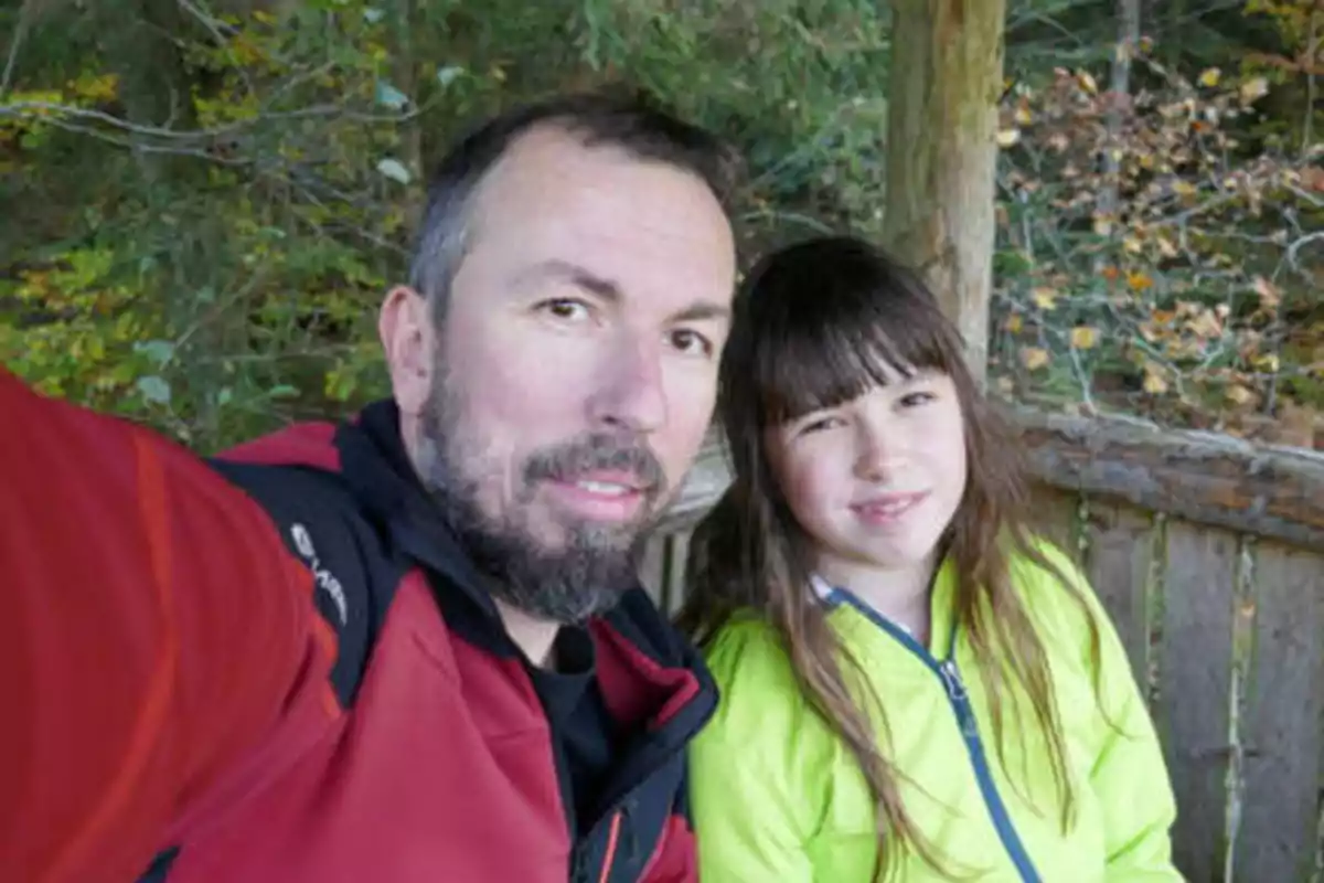 Un hombre y una niña posando juntos al aire libre con árboles de fondo.