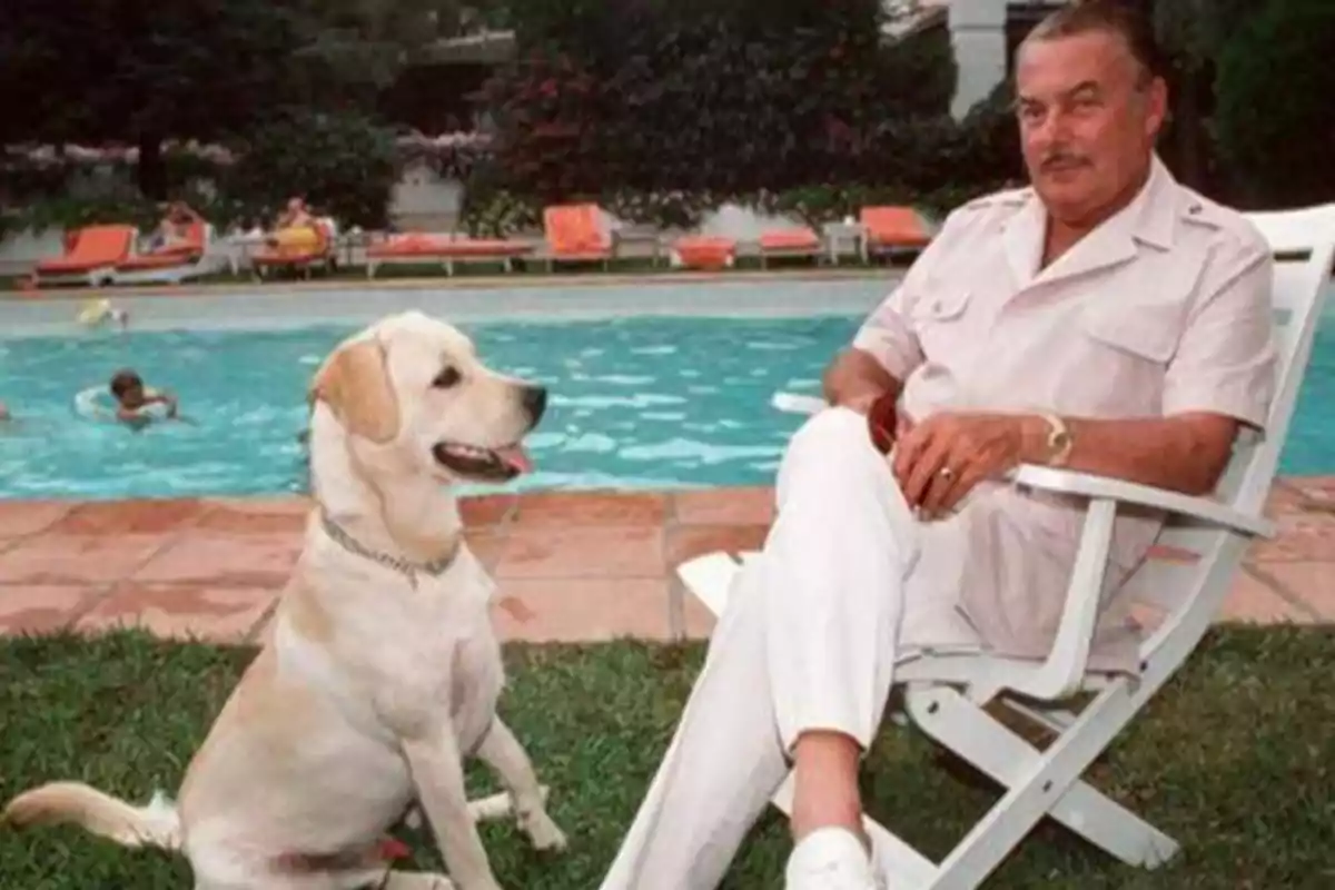 Hombre sentado en una silla junto a una piscina con un perro labrador a su lado.