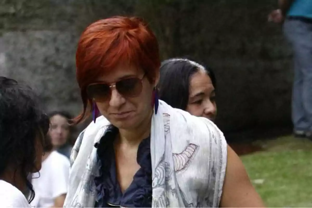 Una persona con cabello corto y rojo lleva gafas de sol y un chal blanco con diseño mientras está al aire libre acompañada de otras personas.