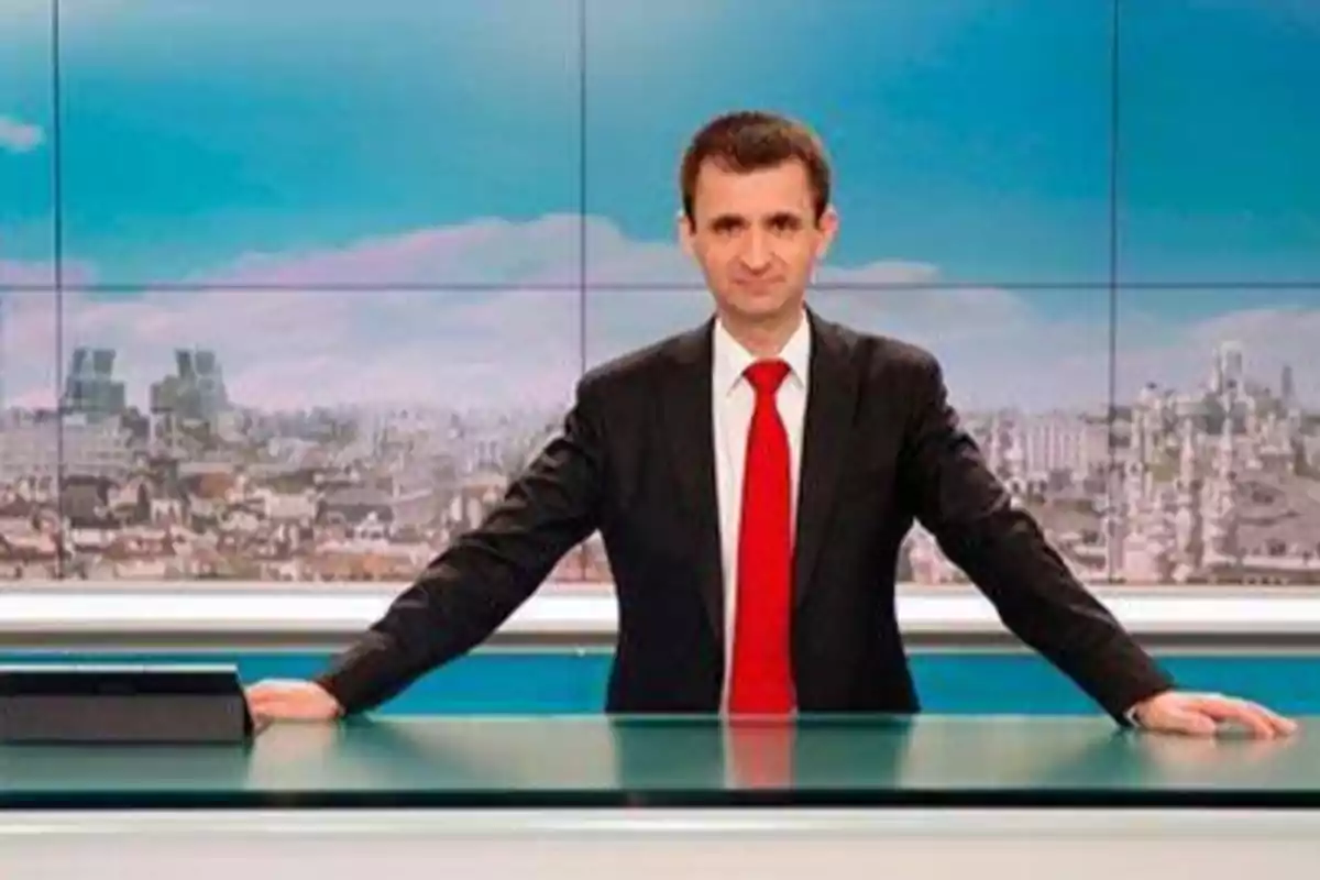 Un hombre con traje y corbata roja está de pie detrás de un escritorio de noticias con una ciudad de fondo.