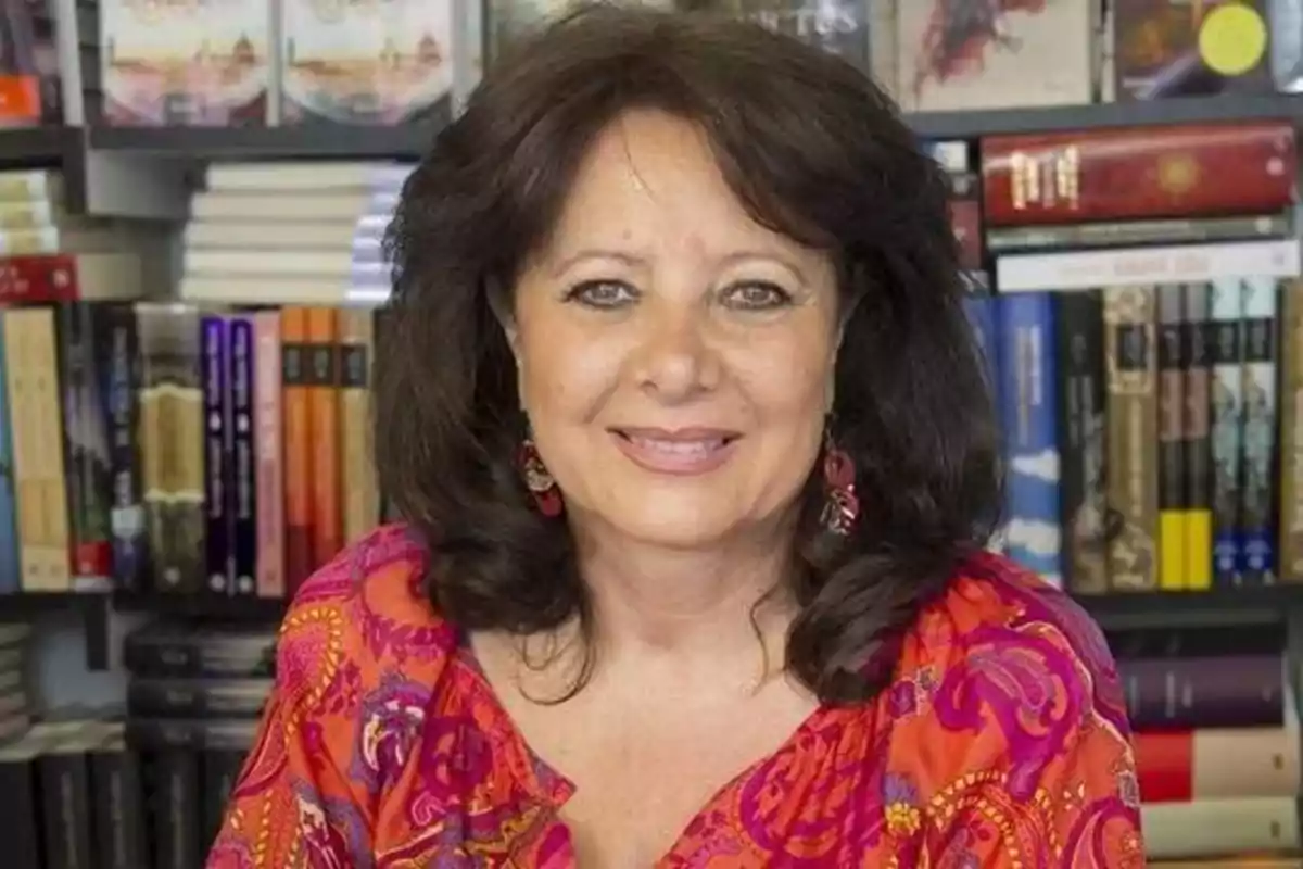 Mujer sonriente con cabello oscuro y blusa colorida, sentada frente a una estantería llena de libros.