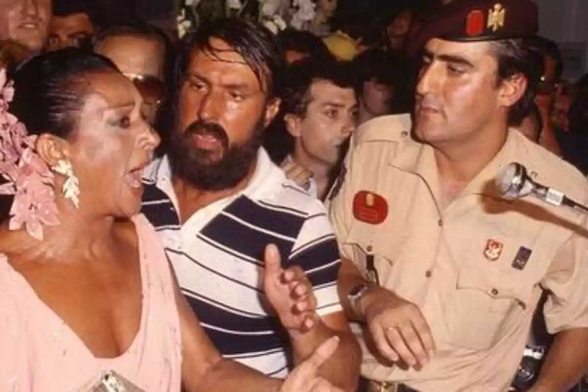 Una mujer con un vestido rosa y adornos florales en el cabello parece estar hablando apasionadamente mientras un hombre con barba y camiseta a rayas y un hombre uniformado la observan en un entorno concurrido.