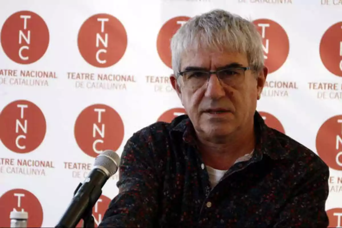 Hombre con gafas y cabello canoso hablando frente a un micrófono con el logo del Teatre Nacional de Catalunya de fondo.