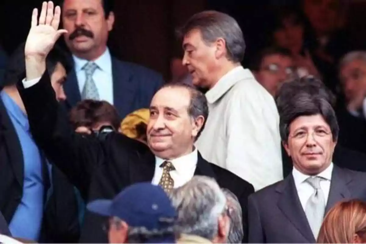 Un hombre de traje oscuro y corbata levanta la mano mientras sonríe, rodeado de otras personas en un evento público.