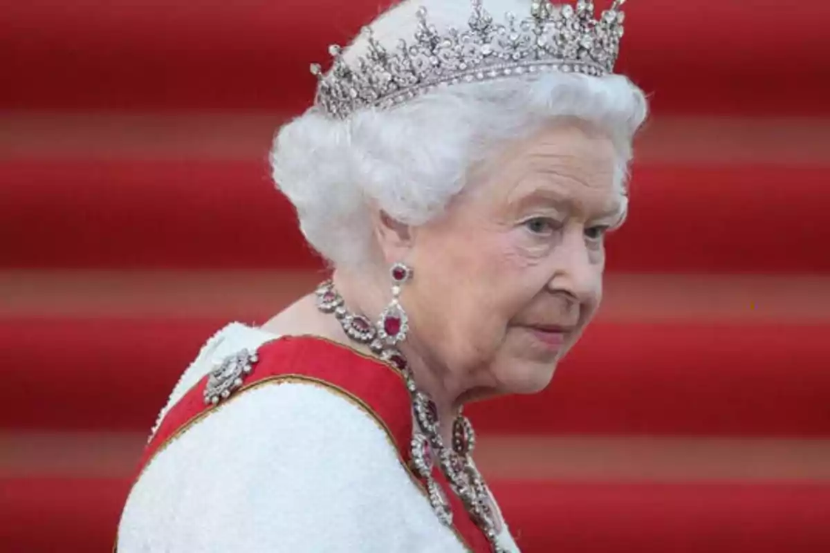 Una mujer mayor con cabello blanco lleva una corona y joyas elegantes, con un fondo de escalones rojos.