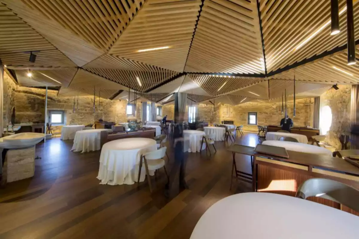 Un elegante restaurante con techo de diseño geométrico de madera, mesas redondas con manteles blancos y paredes de piedra.