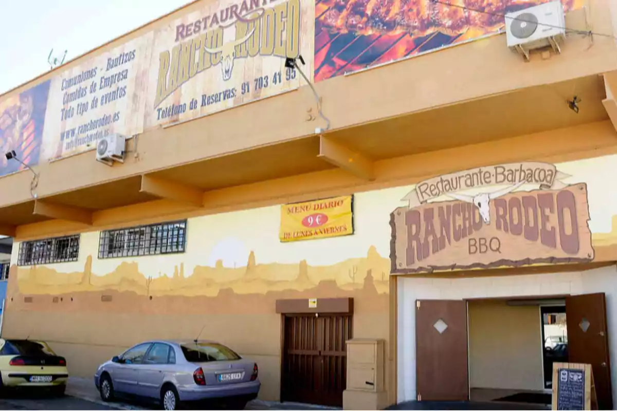 Fachada de un restaurante llamado Rancho Rodeo BBQ con carteles promocionales y dos coches aparcados enfrente