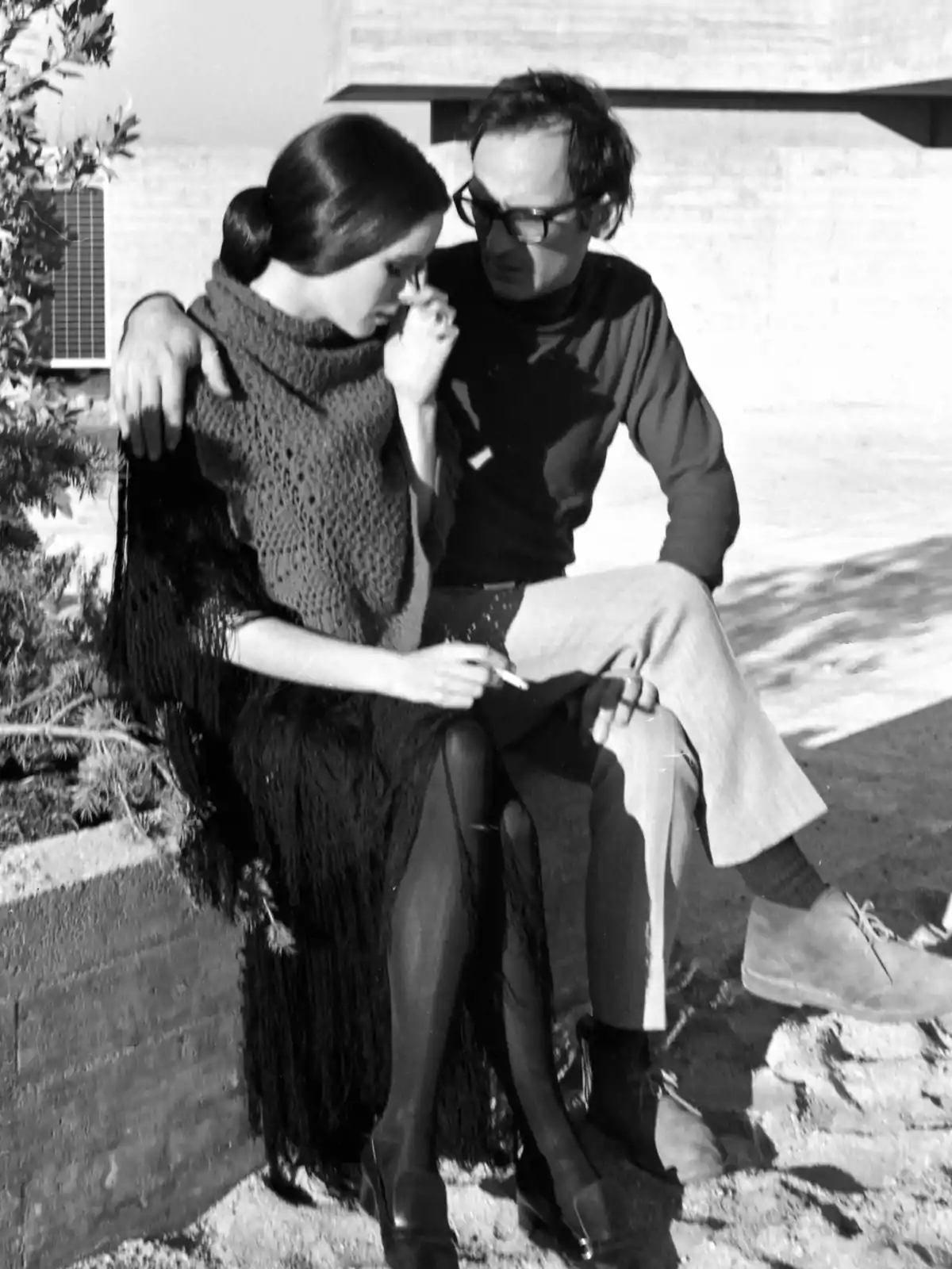 Una mujer y un hombre sentados en un muro de concreto, la mujer está fumando y el hombre la abraza mientras conversan.