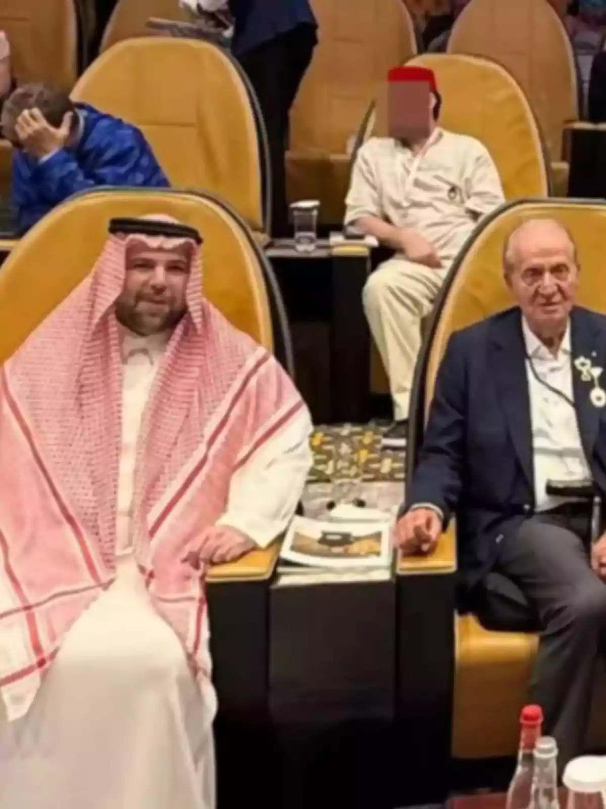 Dos hombres sentados en sillones de color amarillo en un auditorio uno lleva vestimenta tradicional árabe y el otro viste traje oscuro hay personas al fondo Dos hombres sentados en sillones de color amarillo en un auditorio uno lleva vestimenta tradicional árabe y el otro viste traje oscuro hay personas al fondo