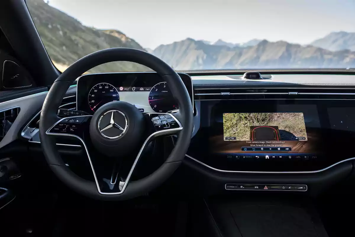 Interior de un Mercedes-Benz E 300 de 4MATIC All-Terrain de lujo con volante y pantallas digitales mostrando información de conducción y cámara de visión trasera, con un paisaje montañoso visible a través del parabrisas. Interior de un Mercedes-Benz E 300 de 4MATIC All-Terrain de lujo con volante y pantallas digitales mostrando información de conducción y cámara de visión trasera, con un paisaje montañoso visible a través del parabrisas.