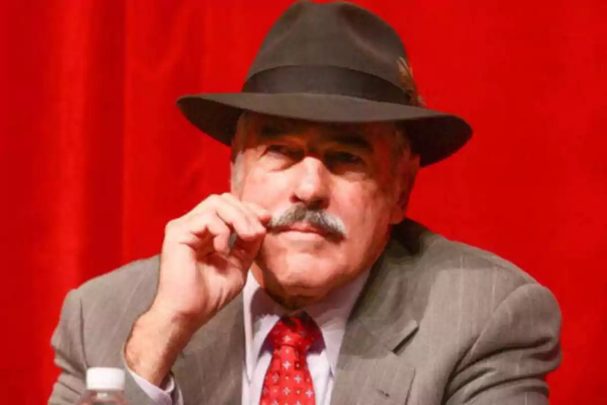 Hombre con sombrero y traje gris sentado frente a un fondo rojo.
