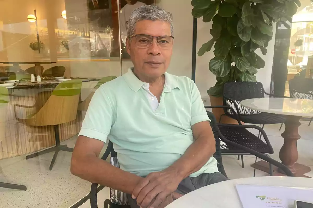 Un hombre con gafas y una camisa polo verde claro está sentado en una mesa en un café moderno con sillas de diseño y plantas decorativas.