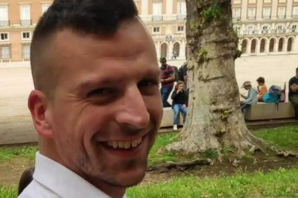 Un hombre sonriente con cabello corto y barba ligera está al aire libre, con un edificio histórico y varias personas sentadas en el fondo.