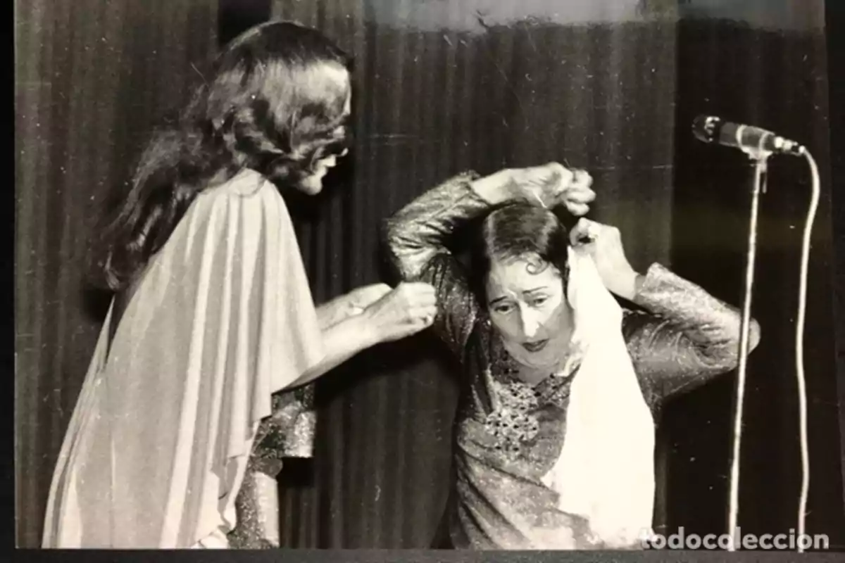 Dos mujeres en un escenario, una ayudando a la otra a colocarse un pañuelo en la cabeza, con un micrófono al fondo.