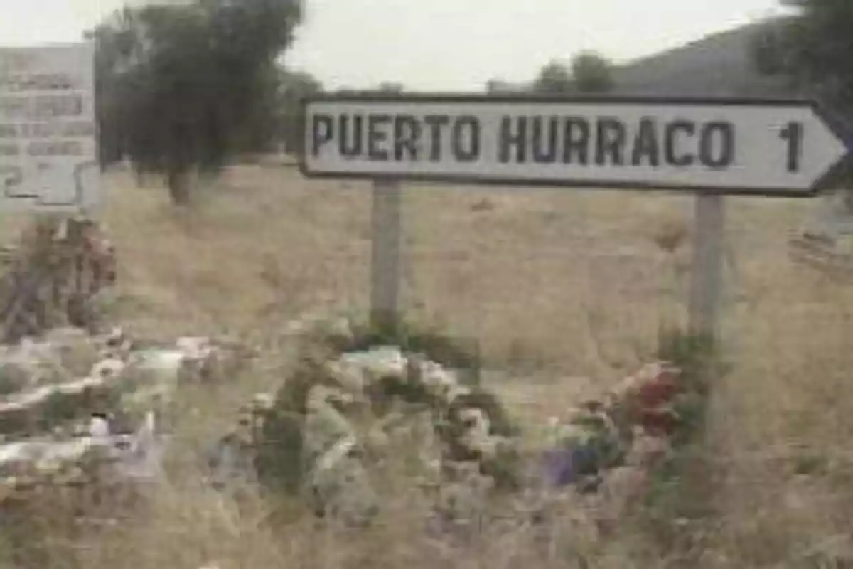 Señal de tráfico que indica la dirección hacia Puerto Hurraco, rodeada de flores y un paisaje rural.