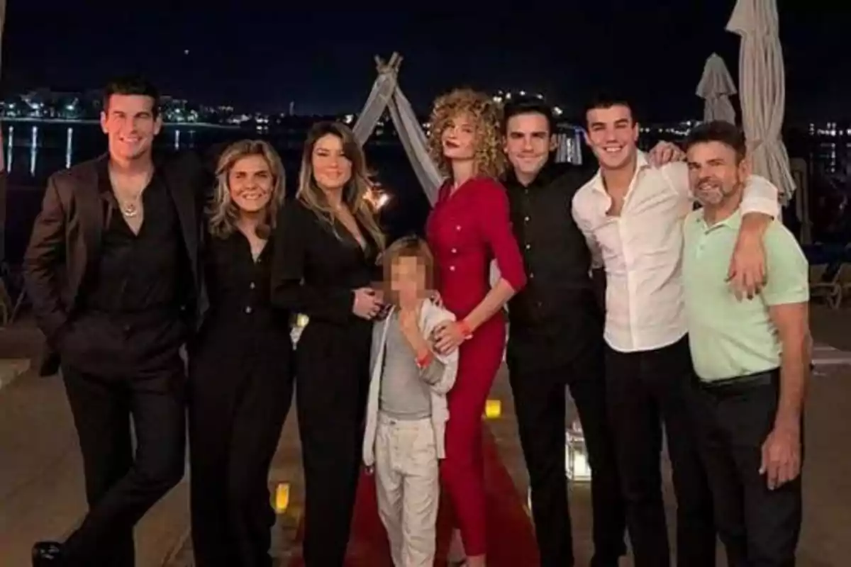 Un grupo de personas posando juntas en una noche al aire libre, con luces de la ciudad y el agua de fondo.