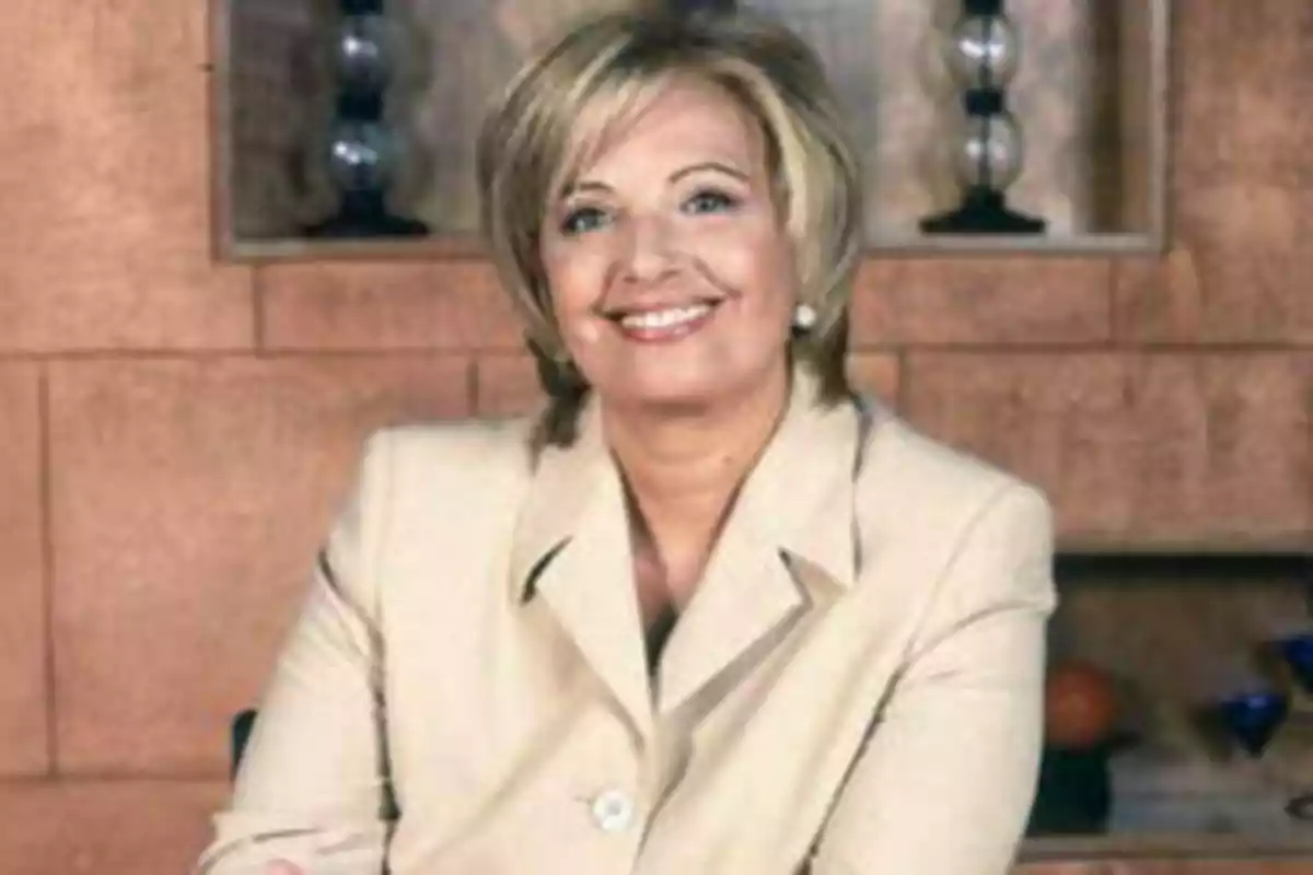 Mujer sonriendo con cabello rubio corto y chaqueta beige, sentada frente a una pared de ladrillos con estanterías decorativas.